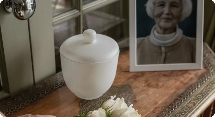 Imagem com um vaso branco, um porta-retrato e flores brancas em um ambiente aconchegante. Refere-se à memória e homenagem.