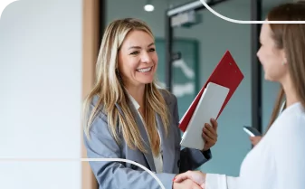 Mulher sorridente com pastas se cumprimentando em ambiente corporativo. Imagem reflete interação profissional positiva e colaboração.