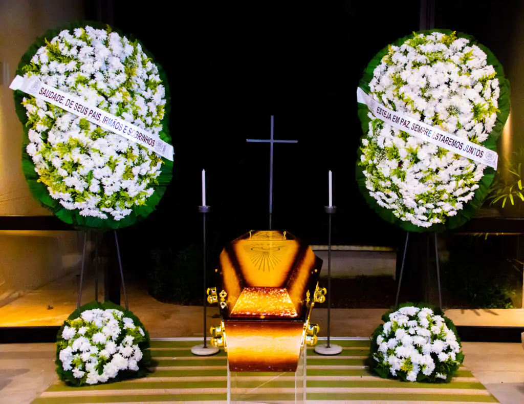 Imagem de uma homenagem póstuma com flores e coroas em um ambiente interno, destacando um caixão dourado. Mensagens de condolências podem ser vistas nas coroas de flores.