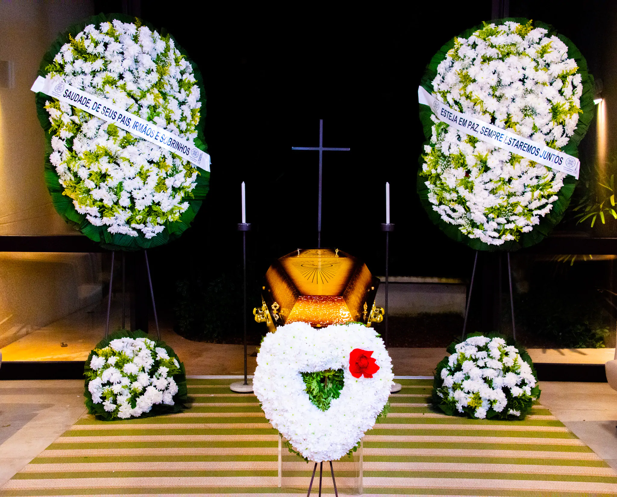 Imagem de uma homenagem póstuma com flores e coroas em um ambiente interno, destacando um caixão dourado. Mensagens de condolências podem ser vistas nas coroas de flores.