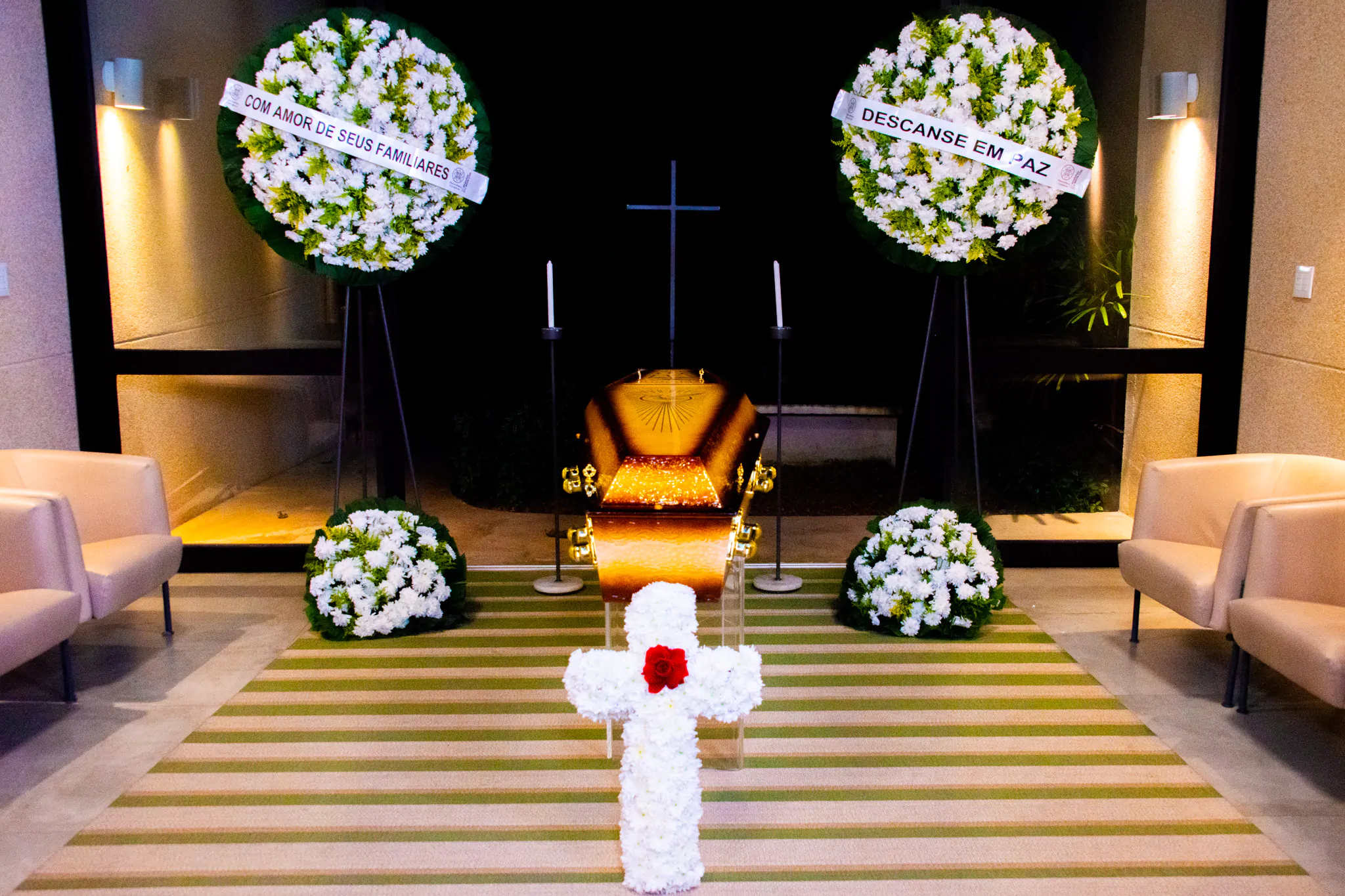 Imagem de uma homenagem póstuma com flores e coroas em um ambiente interno, destacando um caixão dourado. Mensagens de condolências podem ser vistas nas coroas de flores.