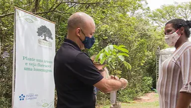 Um homem e uma mulher participando de uma atividade de plantio de árvore, com uma placa ao fundo que diz 'Plante uma árvore e veja florescer uma homenagem'.