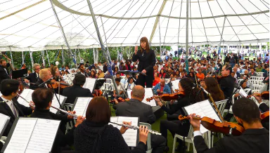Uma orquestra se apresenta sob uma tenda, com uma diretora regendo e um público animado desfrutando da música ao ar livre.