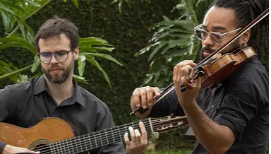 Dois músicos tocam instrumentos em um ambiente natural. Um deles está com um violão e o outro com um violino, criando uma harmoniosa apresentação musical.